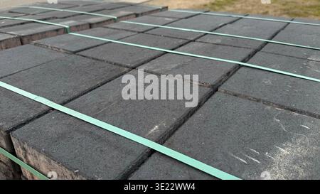 Gestapelte graue Betonpflastersteine, die mit grünen Bändern auf Holzpaletten neben einem Kiesbereich gesichert sind Stockfoto