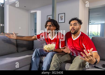 Männliche Freunde sitzen zu Hause auf einem grauen Sofa in roten Trikots, halten Popcorn und reagieren auf das Fernsehen Stockfoto