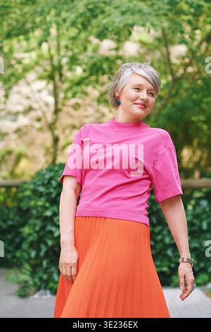 Lächelnde Frau in ihren fünfziger Jahren mit grauen Haaren im grünen Park, in hellrosa Oberteil und orangem Rock, stilvolle reife Frau genießt friedliche Outdoor-L Stockfoto