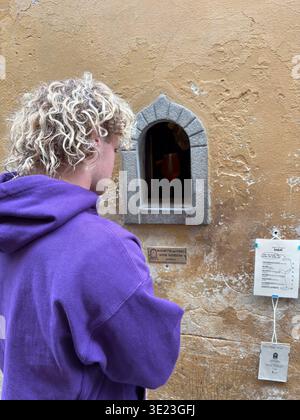 Weinfenster Buchetta del Vino. Florenz, Italien. Junger Mann sammelt Getränke von einem historischen kleinen Service, der in einer strukturierten gelben Wand geöffnet ist. Stockfoto