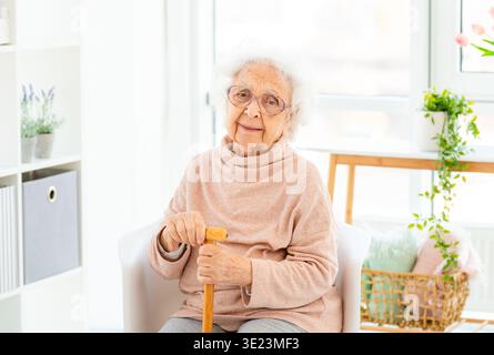 Ältere Frau, die zu Hause sitzt und Einen Stock hält, schaut in Einem hellen, gemütlichen Raum auf die Kamera Stockfoto