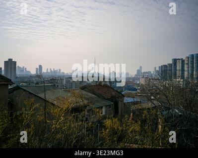Breiter Panoramablick über traditionelle, alte, gekachelte Dächer, der in Richtung einer verdunkelten, fernen modernen Skyline mit einem markanten, hohen Fernseher führt Stockfoto