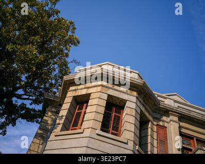 Die Aufwärtsperspektive einer klassischen grauen Steinbauecke mit traditionellen roten Holzfenstern, eingerahmt von grünen Bäumen gegen ein klares blau Stockfoto