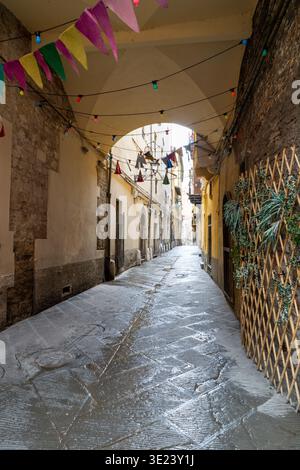 Eine charmante, nasse Kopfsteinpflasterstraße in einer Altstadt, die mit lebhaften Wimpeln und beleuchteten Glühbirnen geschmückt ist und eine festliche Atmosphäre schafft Stockfoto