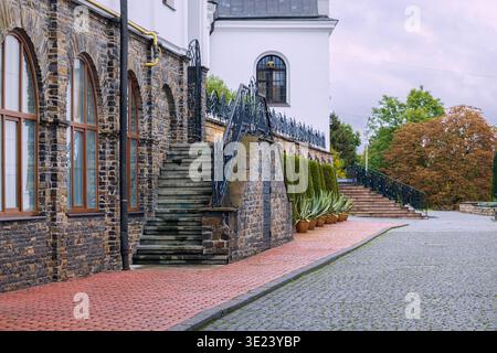 Ein altes Steingebäude mit Bogenfenstern und einer verwitterten Treppe vor einem Hintergrund von üppigem Grün und einem bezaubernden Kopfsteinpflasterweg Stockfoto