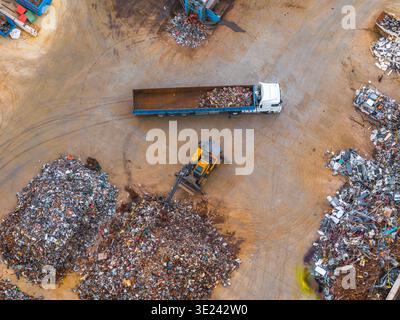Aus der Vogelperspektive einer pulsierenden Recyclinganlage, wo ein gelber Bagger einen weißen LKW zwischen Metallhaufen unter freiem Himmel lädt, Avezzano, Abruzzen, Stockfoto