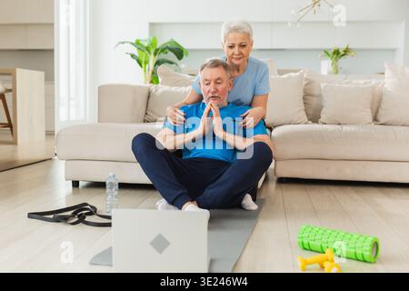 Yoga-Achtsamkeitsmeditation. Älteres erwachsenes Ehepaar, das Yoga mit Online-Unterricht im Laptop praktiziert. Alte Frau, die dem Ehemann hilft, sich zu entspannen. Familie, die zu Hause Atemübungen macht Stockfoto