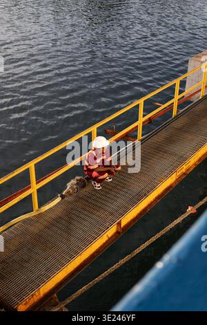 Seefahrer in roter Uniform und Schutzhelm auf einer Metallgangway in einem indonesischen Hafen. Hochwinkel-vertikale Aufnahme für Arbeitssicherheit. Stockfoto