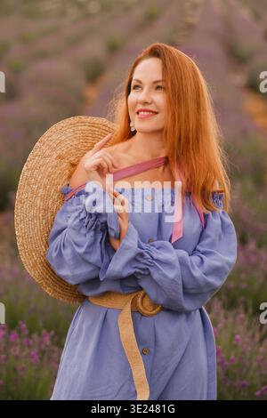 Eine Frau steht in einem Lavendelfeld, trägt ein blaues Kleid mit einem breitkrempigen Strohhut. Sie lächelt und genießt das sonnige Wetter in einer ländlichen Gegend Stockfoto