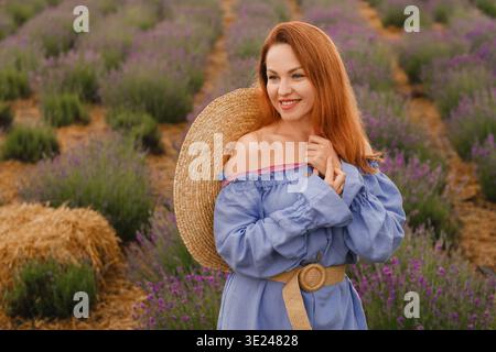 Eine Frau steht in einem Lavendelfeld, trägt ein blaues Kleid mit einem breitkrempigen Strohhut. Sie lächelt und genießt das sonnige Wetter in einer ländlichen Gegend Stockfoto