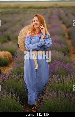 Eine Frau steht in einem Lavendelfeld, trägt ein blaues Kleid mit einem breitkrempigen Strohhut. Sie lächelt und genießt das sonnige Wetter in einer ländlichen Gegend Stockfoto