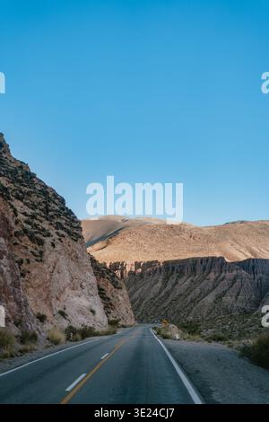 Die Asphaltstraße schlängelt sich durch die zerklüftete Wüstenschlucht. Felsige Klippen bilden den Rahmen für die Reise unter dem klaren blauen Himmel. Sonnenlicht leuchtet warm auf fernen Bergrücken Stockfoto