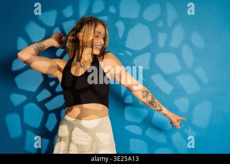 Frau mit Dreadlocks und Tattoos, die sich vor einer blauen Wand mit abstrakten Lichtmustern posiert Stockfoto