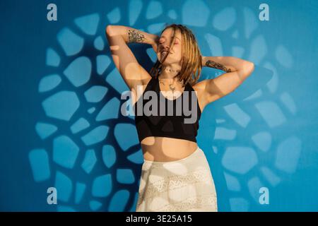 Frau mit Dreadlocks und Tattoos, die sich vor blauem Hintergrund mit Schattenmustern posiert Stockfoto