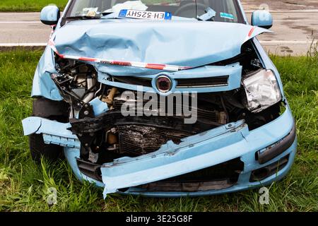 Vorderansicht eines schwer beschädigten hellblauen Fiat Panda auf einer grasbewachsenen Straße, mit einer zerquetschten Motorhaube, freiliegendem Kühler und italienischem Kennzeichen Stockfoto