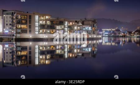 Nordischer Hafen lebendige Tradition beleuchtete Wohnblöcke entlang des Flusses in Porsgrunn Norwegen spiegeln sich im stillen Wasser mit strukturierten Balkonen und Stockfoto