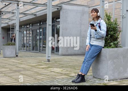 Asiatische Frau 20s sitzt auf Pflanzgefäß in plaza, Denim, Kopfhörer, Tasche, hält Telefon, Kopierbereich Stockfoto