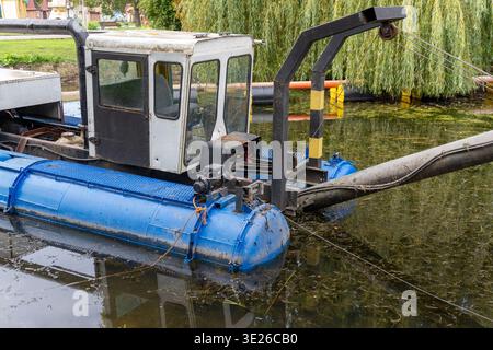 Eine Maschine arbeitet im Wasser, um am Nachmittag in einem Park Schmutz zu sammeln. Bäume und Bänke umgeben die Gegend, während einige Leute den Weg entlang laufen. Stockfoto