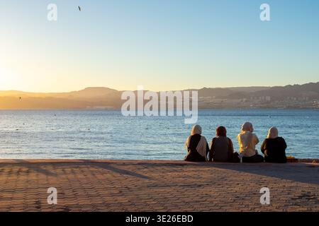 Rückansicht von drei muslimischen Frauen im Hijab, die am Strand sitzen und den Sonnenuntergang in Aqaba, Jordanien, beobachten. Rotes Meer und Blick auf Israel Stockfoto