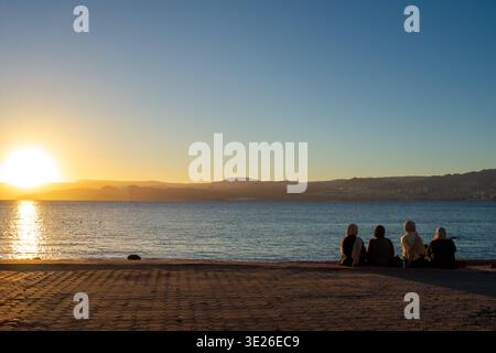 Rückansicht von drei muslimischen Frauen im Hijab, die am Strand sitzen und den Sonnenuntergang in Aqaba, Jordanien, beobachten. Rotes Meer und Blick auf Israel Stockfoto