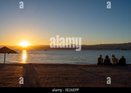 Rückansicht von drei muslimischen Frauen im Hijab, die am Strand sitzen und den Sonnenuntergang in Aqaba, Jordanien, beobachten. Rotes Meer und Blick auf Israel Stockfoto