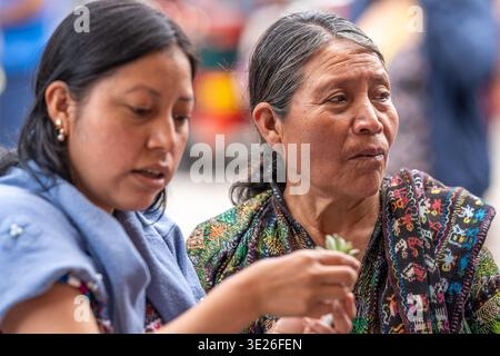 Festival de Flores - Antigua Guatemala - zwei indigene guatemaltekische Frauen verbringen einen ruhigen Moment, um bunte gewebte Textilien auf einem Markt im Freien zu untersuchen. Stockfoto