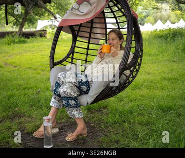 Frau, die sich im hängenden Eierstuhl entspannt und unter einem Baum Tee trinkt, in der englischen Landschaft Stockfoto