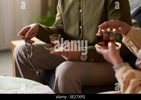 Muslimischer Mann mit Gebetskugeln und einem Glas Tee sitzend mit muslimischer Frau, die im Wohnzimmer Tee teilt und in entspannter Unterhaltung teilnimmt Stockfoto