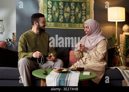 Junger muslimischer Mann, der Gebetsperlen hält und Tee trinkt, mit einer jungen muslimischen Frau, die einen Hijab trägt und lächelt, während sie zusammen auf dem Sofa sitzt und Snacks teilt Stockfoto