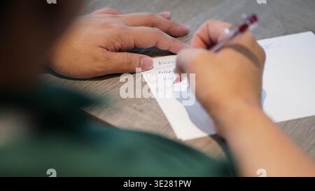 Nahaufnahme einer Person, die eine persönliche Liste mit einem roten Stift auf Papier schreibt, die Hand auf einer Holzoberfläche aufliegt. Stockfoto