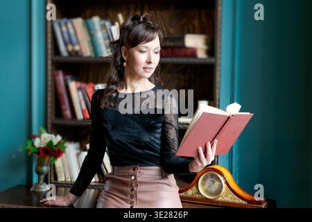 Mittlere Aufnahme einer eleganten Frau in einer schwarzen Spitzenbluse, die in ein offenes rotes Buch schaut. Klassischer Vintage-Bibliothekshintergrund mit Bücherregalen, einer Uhr, einer Stockfoto