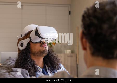 Verschiedene männliche Freunde beobachten afroamerikanischen Mann mit weißem VR-Headset auf dem Sofa zu Hause Stockfoto