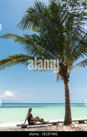 Junge Frau, die auf einer Holzbank unter Palmen sitzt, mit Blick auf das türkisfarbene Meer, tropische Strandreise-Lifestyle-Szene Stockfoto