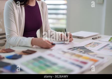 Fokussierte Erwachsene asiatische Frau zeichnet mit Bleistift Design-Pläne und wählt lebendige Farbmuster auf einem modernen Schreibtisch für kreative Projekte Stockfoto
