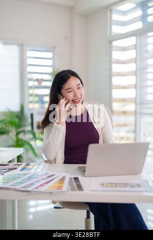 Junge asiatische Frau lächelt mit ihrem Smartphone im modernen Home Office. Sie verwendet einen Laptop, während sie geschäftliche oder Designideen aus der Ferne diskutiert. Dieses Bild Co Stockfoto
