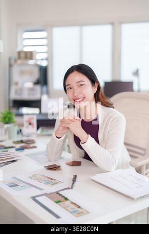 Asiatische Frau, die selbstbewusst am Schreibtisch lächelt. Architekturpläne, Materialmuster, Entwurfspläne für Wohnprojekte auf dem Tisch. Kreative Arbeit Stockfoto