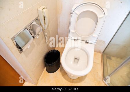 Weiße Toilette mit offenem Deckel im Hotelbadezimmer mit beigefarbenen Fliesen, Telefon zur Wandmontage und schwarzem Müll Stockfoto