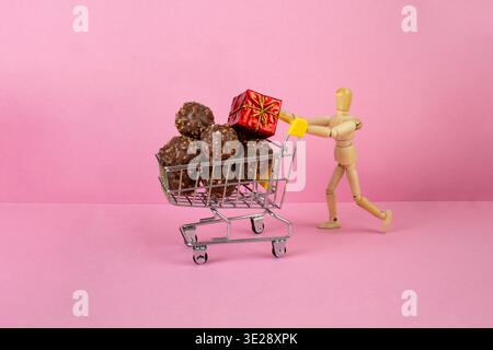 Hölzerne Schaufensterpuppe schiebt einen Miniatur-Einkaufswagen gefüllt mit Schokoladenkugeln mit Walnüssen auf rosa Hintergrund, süßes Geschenk Verkaufskonzept Stockfoto