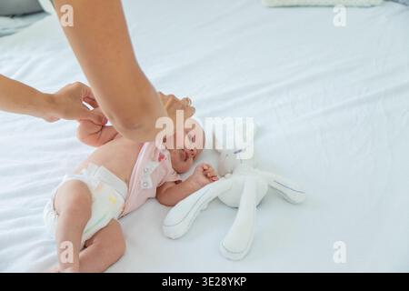 Mutter, die Babykleidung auf weißem Bett wechselt, Kind in Windel in der Nähe von Kuschelspielzeug Stockfoto