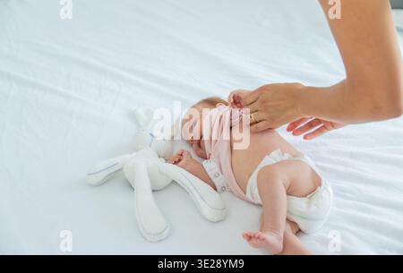 Mutter, die Babykleidung auf weißem Bett wechselt, Kind in Windel in der Nähe von Kuschelspielzeug Stockfoto