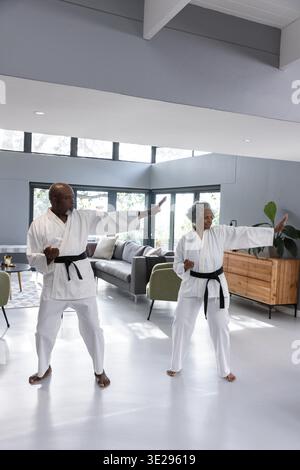 Diverse Seniorenpaare üben Karatepaare im Wohnzimmer mit weißem gis und schwarzem Gürtel. Kampfkunst, Fitness, Wellness, Heimtraining, Kraft, aktives Altern, moderner Innenraum || Modell veröffentlicht Stockfoto