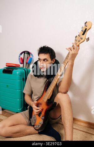 Porträt eines jungen Mannes im Nackenkissen sitzt neben einem türkisfarbenen Koffer in einem Flughafenterminal, spielt Bassgitarre und trägt Kopfhörer entspannt Stockfoto