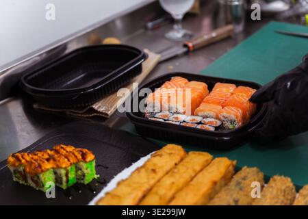 Verschiedene Sushi- Und Gebratene Rolls Zum Mitnehmen Im Black Container, Koch Bereitet Ausgewogen Vor Stockfoto
