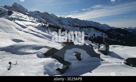 Blick aus der Vogelperspektive auf schneebedeckte Chalets, eingebettet in die hoch aufragenden, schneebedeckten Berge unter dem hellen Himmel, Le Grand-Bornand, Auvergne-Rhône-Alpes, F Stockfoto