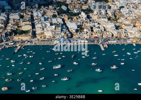 Blick aus der Vogelperspektive auf den ruhigen Hafen mit farbenfrohen Booten, die im Kontrast zu den alten Gebäuden entlang der Küste stehen, Marsaxlokk, Malta. Stockfoto