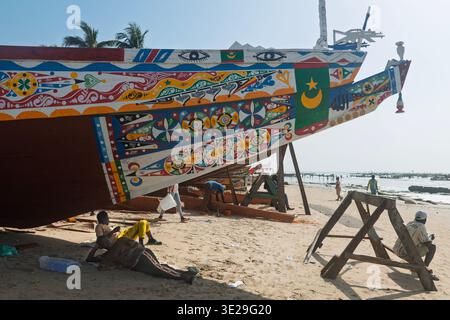 Fischer, die sich im Schatten eines bunt bemalten Bugs einer Piroge am Strand von Kafountine, Casamance, Senegal entspannen. Stockfoto