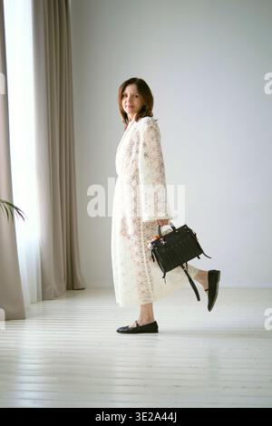 Frau in einem weißen Spitzenkleid hält eine schwarze Handtasche, während sie in einem hellen steht Stockfoto