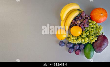 Eine bunte Auswahl an Früchten mit Kürbis auf grauem Hintergrund. Eine lebendige Präsentation von frischem Obst, einschließlich Bananen, Trauben, Orangen, Pflaumen, Avocado Stockfoto