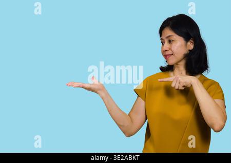 Frau lächelt und selbstbewusst, eine Hand offen für das Produkt und die andere Hand zeigt auf Ihr Produkt. Isoliert auf blauem Hintergrund. Schönes asiatisches Modell. Stockfoto