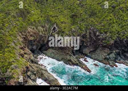 Blick von der Drohne auf Sharkies Beach mit einer zerklüfteten felsigen Küste und einer mit Bäumen bedeckten Bergkulisse. Stockfoto